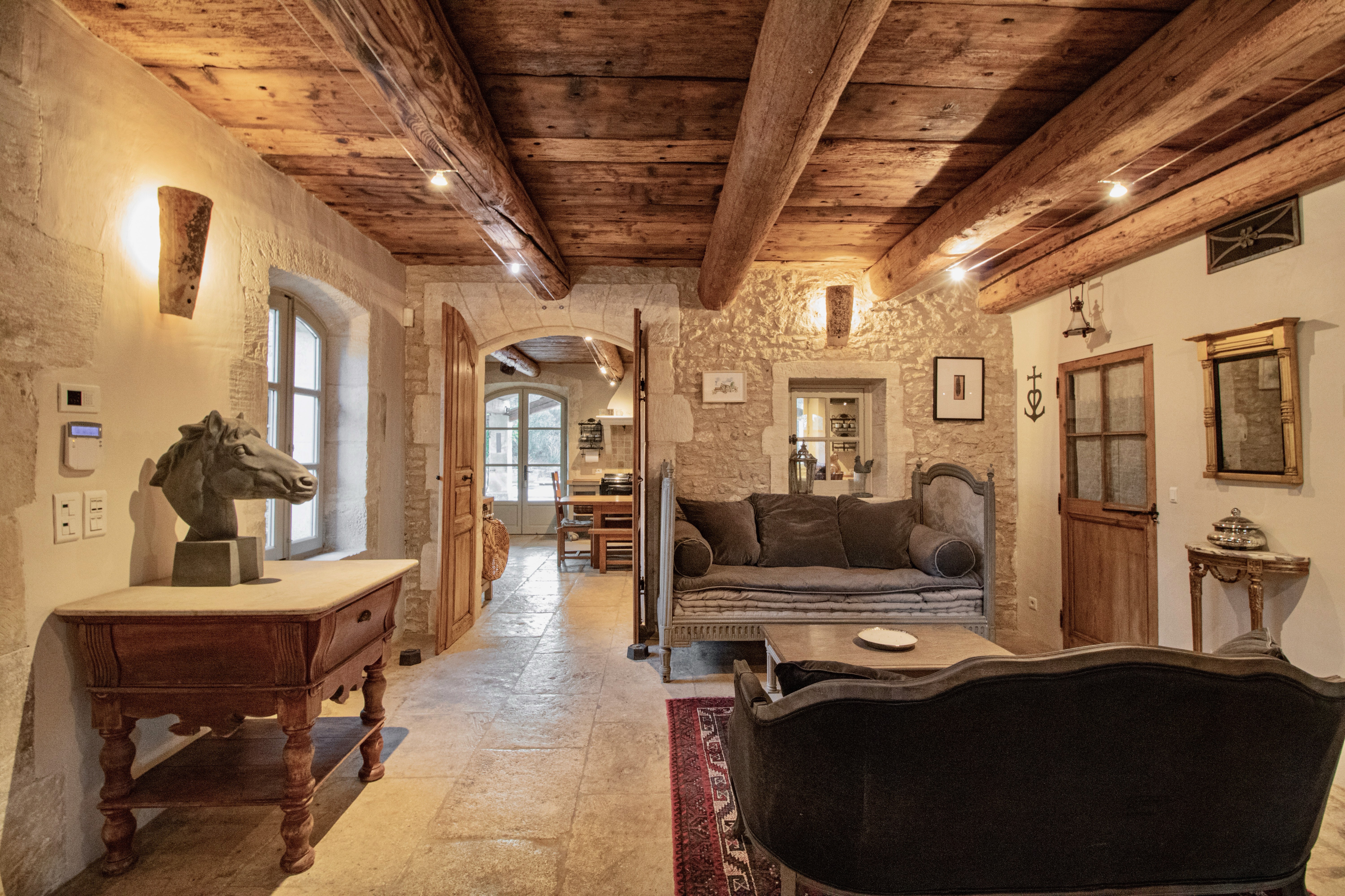 Interior of a rustic living room with exposed wooden beams, stone walls, and vintage furniture including a sofa, wooden tables, and a horse head sculpture.